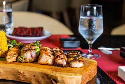 Grilled meats and vegetables on a wooden board, with a glass of water and red napkins on a table.