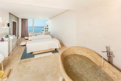 Luxurious spa room with two massage tables, a stone bathtub, and ocean view through large windows, featuring calming neutral tones.