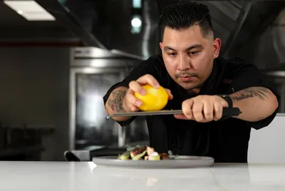 Chef zesting a lemon over a plated dish in a professional kitchen, focused and wearing a black shirt.