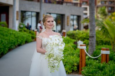 Bride in a white gown holding a cascading bouquet of white flowers, walking along a lush garden path.