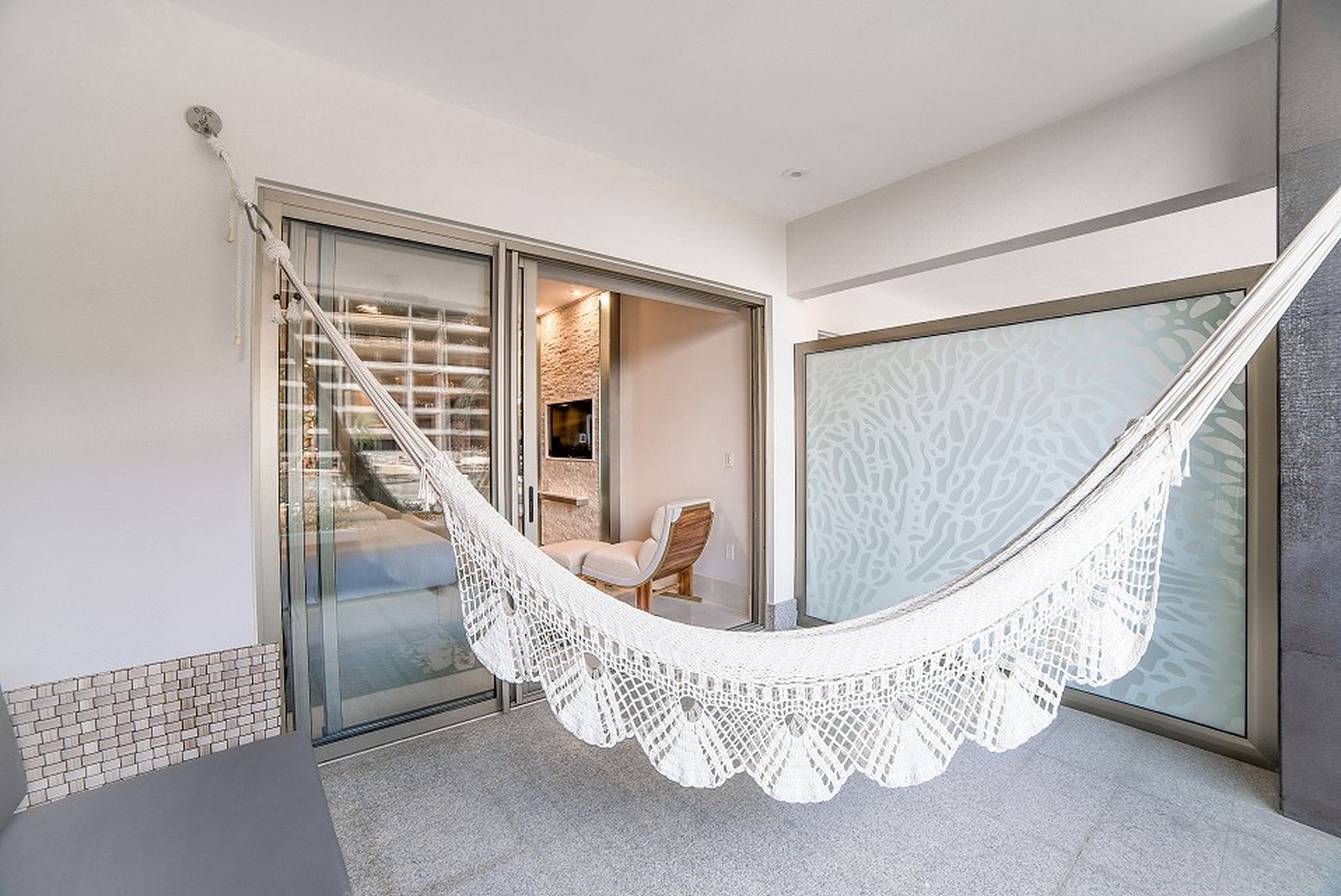 A white hammock hangs in a modern, minimalist room with sliding glass doors, revealing a cozy interior with a wooden chair and textured walls.