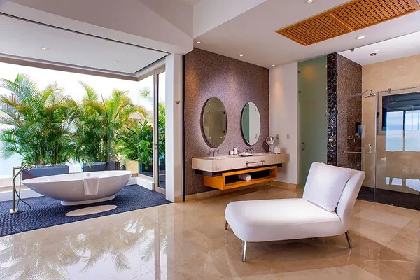 Luxurious bathroom with a freestanding tub, double vanity, round mirrors, white chaise lounge, and large windows overlooking greenery.