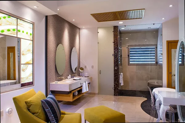Modern bathroom with dual sinks, round mirrors, a walk-in shower, and yellow seating. Stained glass window adds an artistic touch.