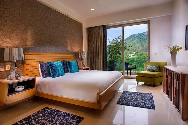 Modern bedroom with a large bed, blue pillows, armchair, and a view of lush green hills through sliding glass doors. Elegant decor and lighting.