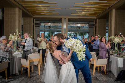 A newlywed couple shares a kiss in an elegant reception hall, surrounded by applauding guests and floral arrangements.
