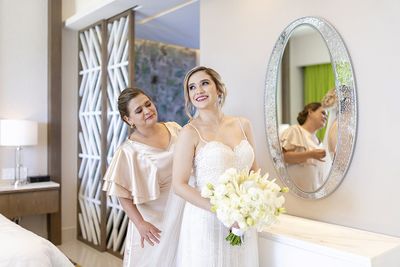 Bride in a white gown holding a bouquet, smiling with a woman in a peach dress adjusting her veil in a bright, elegant room.