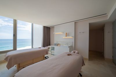 A serene spa room with two massage tables, large window with ocean view, and soft lighting, creating a tranquil atmosphere.