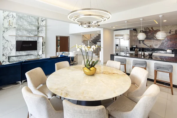 Modern open-plan living area with a round marble dining table, plush chairs, blue sofas, a kitchen island, and elegant lighting fixtures.