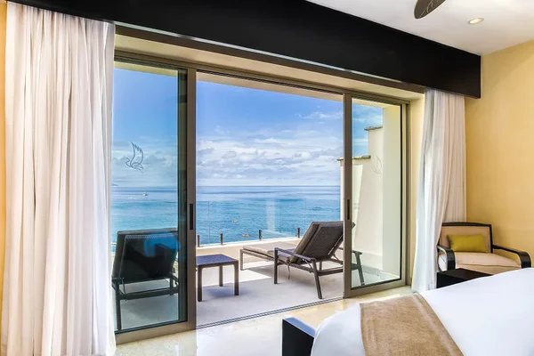 A hotel room with a bed, armchair, and open sliding doors leading to a balcony with lounge chairs, overlooking the ocean under a blue sky.