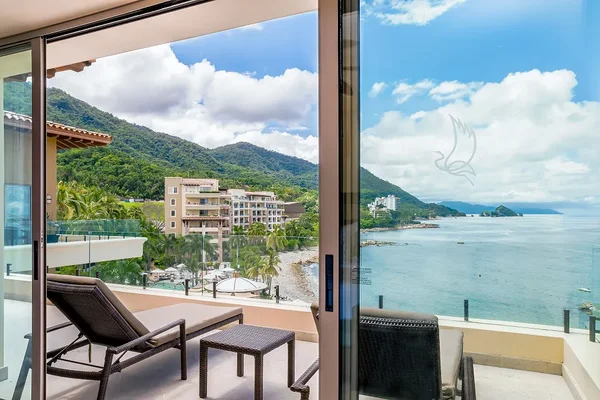 Balcony view of a coastal resort with lounge chairs, overlooking the ocean, mountains, and buildings under a partly cloudy sky.