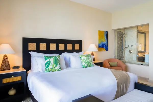 Modern bedroom with a large bed, white linens, tropical-themed pillows, bedside lamps, and a view of a bathroom through a glass partition.