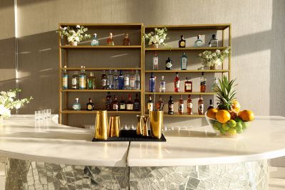 Elegant bar with a variety of spirits on display shelves, gold accents, and a bowl of fresh fruit on a sleek counter.