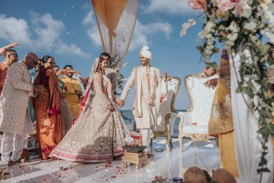 A couple in traditional attire celebrates their wedding on a decorated outdoor platform, surrounded by family under a clear blue sky.
