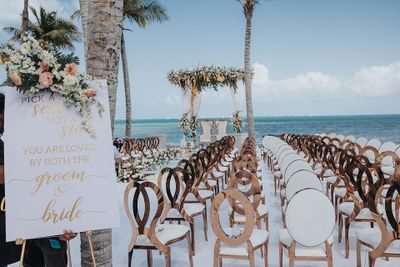 Beachfront wedding setup with elegant chairs, floral arch, and a sign reading, "Pick a seat, not a side. You are loved by both the groom & bride."