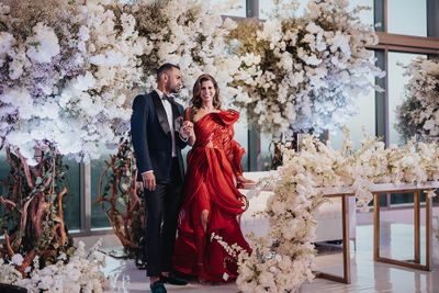 A couple elegantly dressed in formal attire stands amidst lavish floral arrangements at an upscale event.