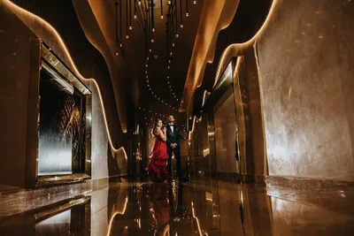A couple elegantly dressed, standing in a luxurious, warmly lit hallway with reflective floors and modern design elements.