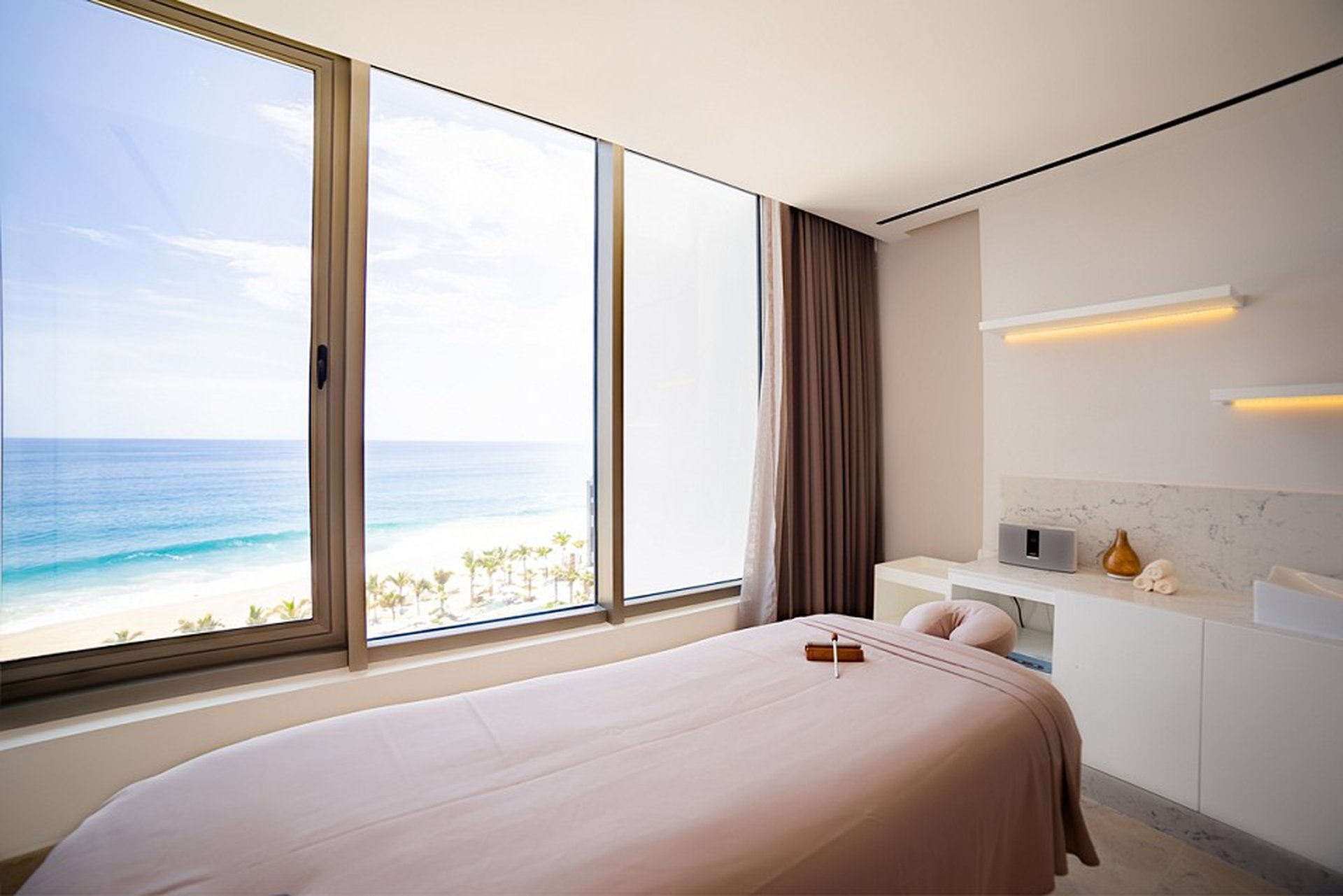 Massage room with a bed facing large windows offering an ocean view. The room has soft lighting, a small counter, and a serene atmosphere.