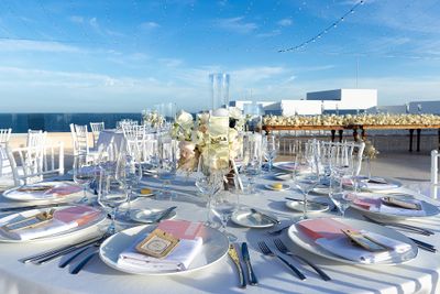 Elegant outdoor wedding table setting with white chairs, glassware, floral centerpieces, and string lights under a clear blue sky by the ocean.