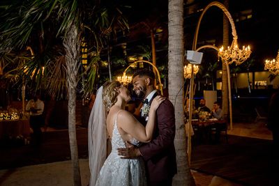 A couple dances joyfully under elegant chandeliers on a moonlit beach, surrounded by palm trees.