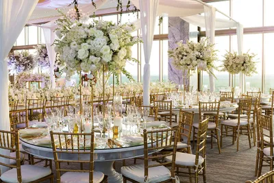 Elegant wedding reception with round tables, gold chairs, and tall floral centerpieces in a sunlit, draped venue.