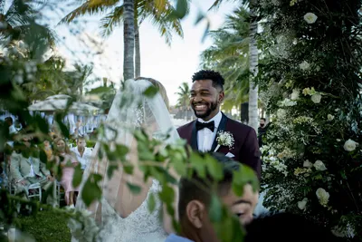 Bride and groom smiling during an outdoor wedding ceremony, surrounded by lush greenery and palm trees.