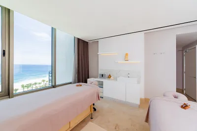 Bright spa room with two massage tables, overlooking a scenic ocean view through large windows. Neutral tones and minimalist decor create a serene atmosphere.