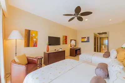 Bright hotel room with two beds, decorative pillows, a wall-mounted TV, armchair, and modern art. Ceiling fan and soft lighting enhance coziness.