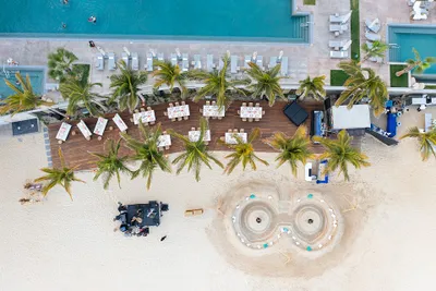 Aerial view of a beachside resort with palm trees, a pool, and sandy areas. Tables are set up on a wooden deck near the water.