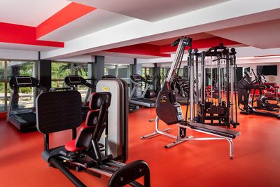 Modern gym with red and white decor, featuring treadmills, weight machines, and large windows with a view of greenery outside.