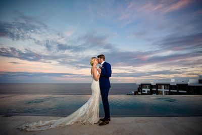 A couple in wedding attire embraces by an infinity pool, with a colorful sunset sky and ocean in the background.