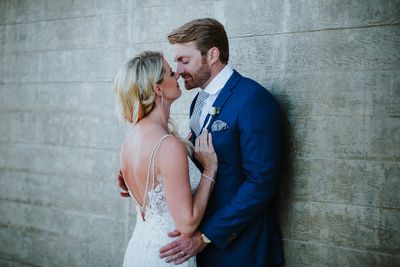 A couple in wedding attire embraces against a textured wall. The bride wears a lace dress, and the groom is in a blue suit.