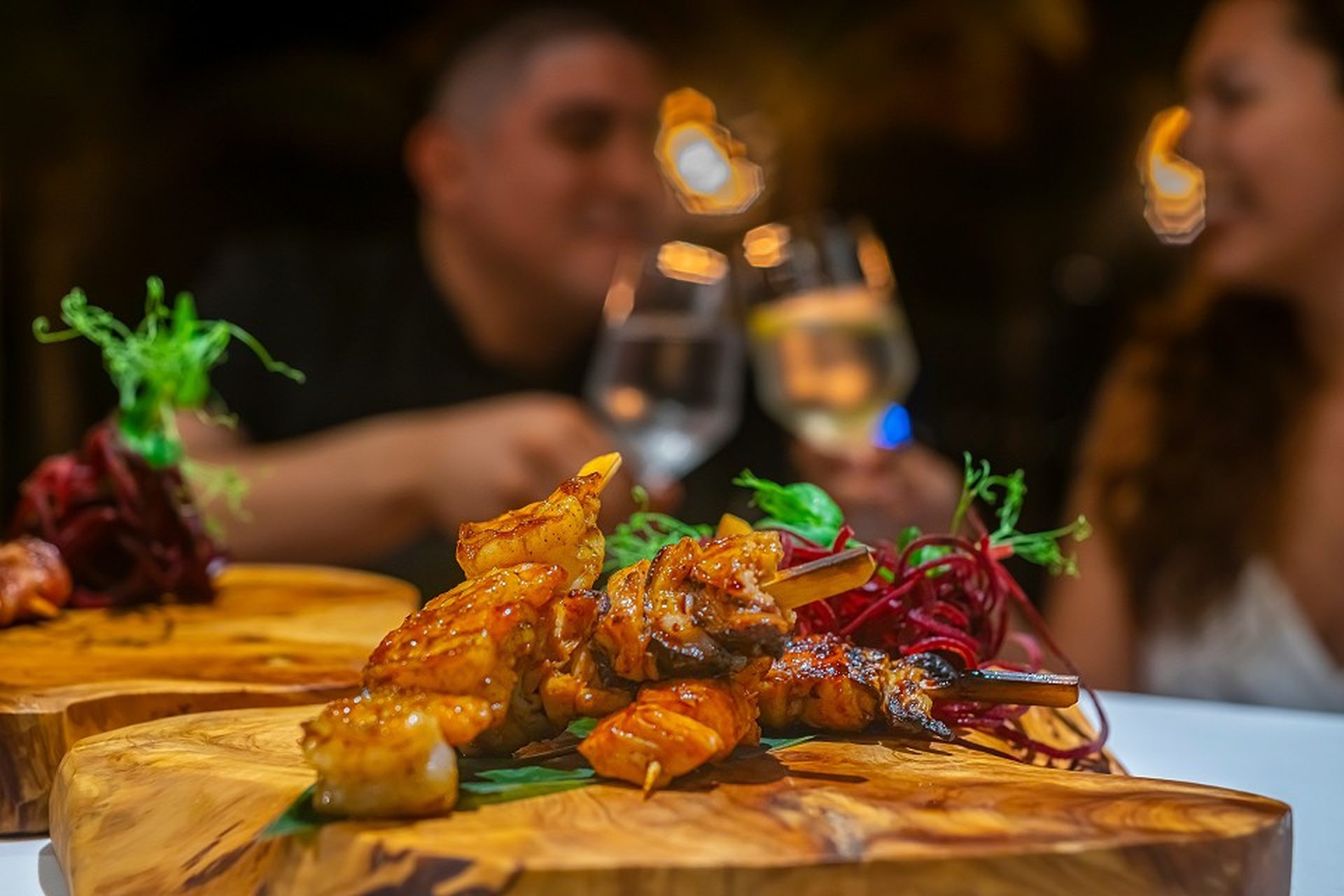 Grilled shrimp skewers on wooden platters with a blurred couple toasting with wine glasses in the background.