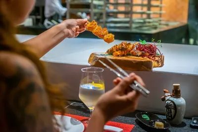 Person holding a skewer of grilled meat over a wooden platter, with a glass of drink and chopsticks in the foreground.