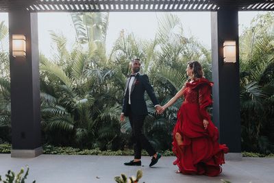 A couple elegantly dressed, holding hands, walking under a pergola with lush greenery in the background.