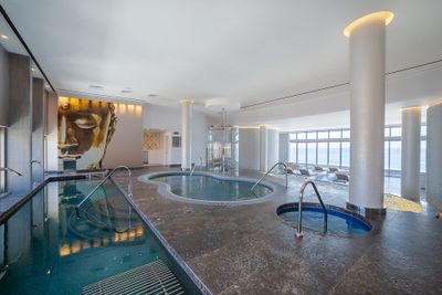Luxurious spa room with a large mural of a serene face, featuring two pools, lounge chairs, and large windows offering ocean views.