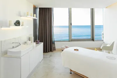 A serene spa room with a massage table, large windows offering an ocean view, white decor, and soft lighting.