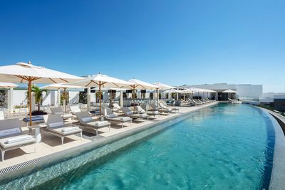 Rooftop infinity pool with sun loungers and umbrellas under a clear blue sky, offering a luxurious and serene atmosphere.