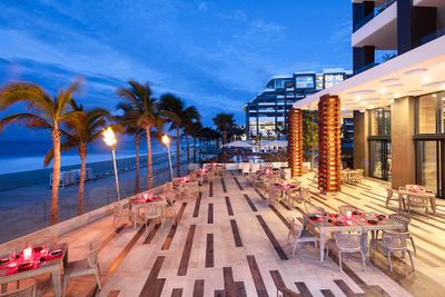 Outdoor restaurant by the beach at dusk, with tables set for dining, palm trees, and modern architecture in the background.