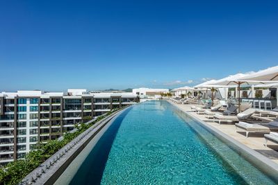 Rooftop infinity pool with lounge chairs and umbrellas, overlooking modern buildings under a clear blue sky.