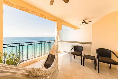 Balcony overlooking the ocean with a hammock, two black chairs, and a ceiling fan under a covered area.