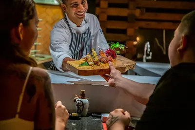 Chef presenting a wooden platter with artfully arranged food to two diners at a restaurant counter.