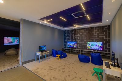 Gaming room with three screens, blue bean bags, and geometric wall pattern. A TV in the adjacent room shows a cartoon.