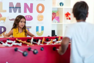 Two children playing foosball in a colorful room with alphabet wall decor. The girl wears a yellow shirt, and the boy is in a white shirt.