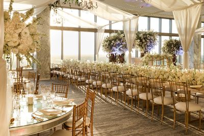 Elegant wedding reception with gold chairs, white floral arrangements, and floor-to-ceiling windows overlooking a sunset.