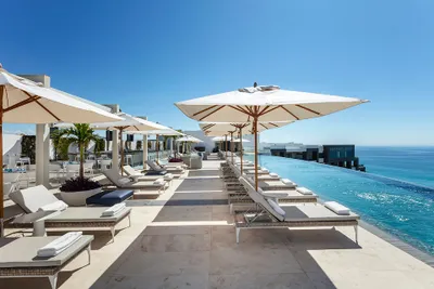 Rooftop infinity pool with sun loungers and umbrellas overlooking the ocean on a clear, sunny day.