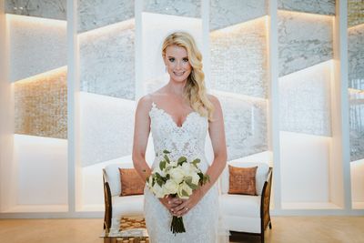 Bride in a lace wedding dress holding a bouquet of white roses, standing in a modern room with a textured wall and soft lighting.