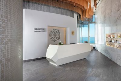 Modern hotel lobby with a sleek white reception desk, geometric wall art, and ocean view. Elegant lighting and display shelves enhance the ambiance.