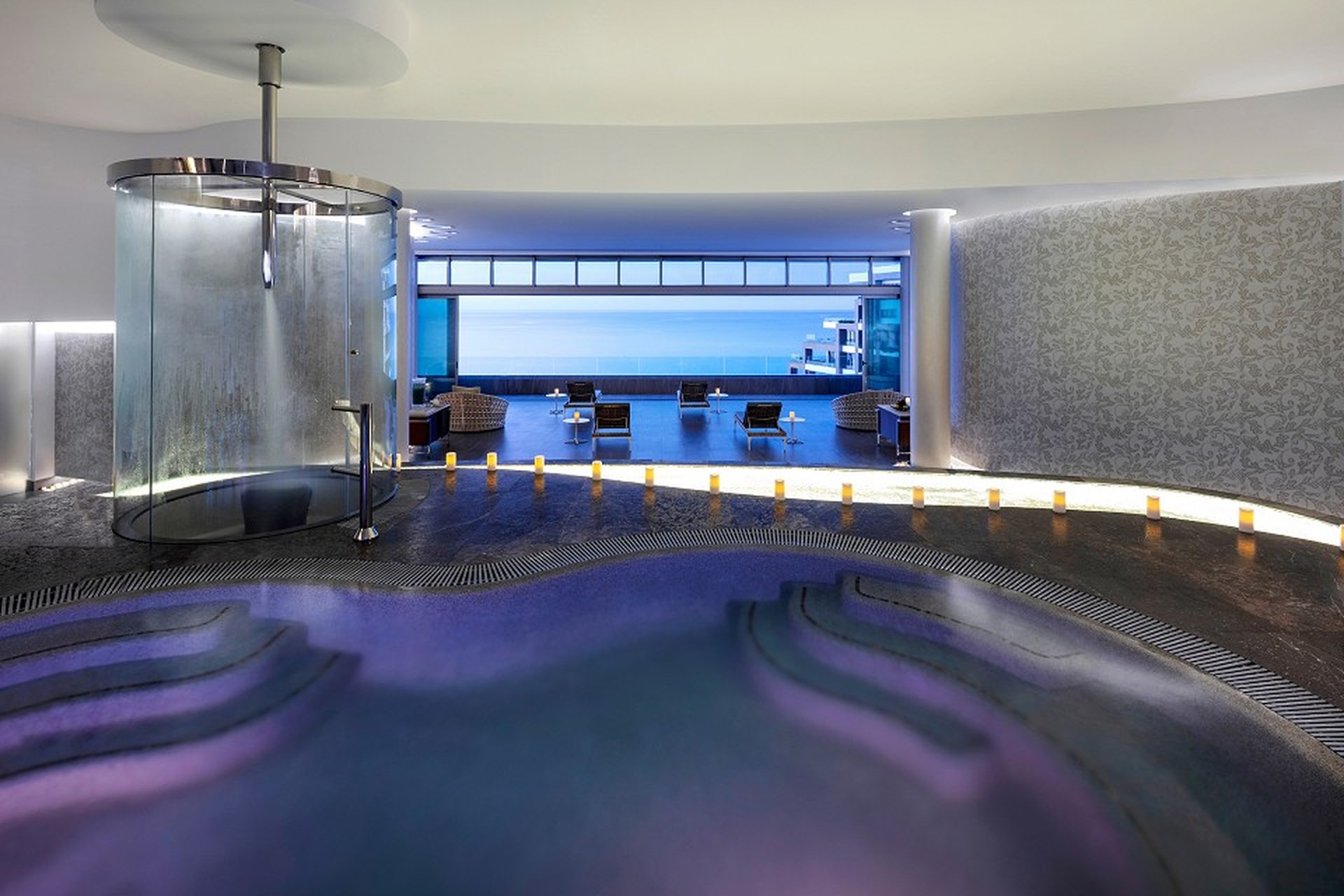 Luxurious spa interior with a circular shower, ambient lighting, and a view of an ocean through large windows. Candles line the path.
