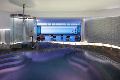 Luxurious spa interior with a circular shower, ambient lighting, and a view of an ocean through large windows. Candles line the path.