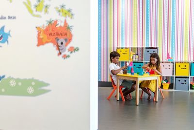 Two children sitting at a small table playing with toys in a colorful room with striped wallpaper and cubby shelves. A map of Australia is partially visible.